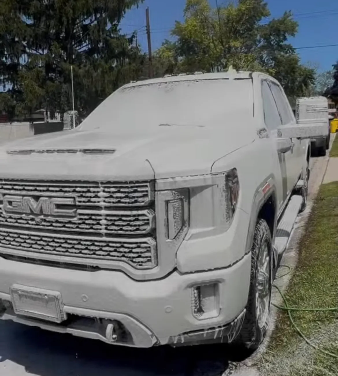 A GMC Sierra Denali covered in a thick layer of snow foam during an exterior wash on a sunny day.